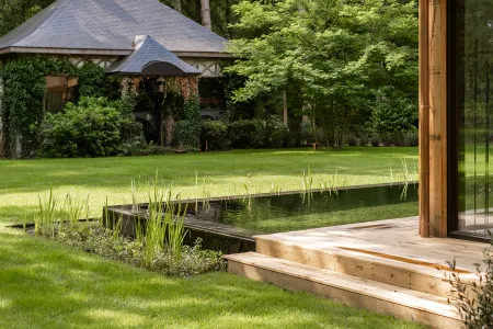 Zwemvijver met rietplanten voor de houten villa, met zicht op een klassiek bijgebouw in Bonheiden