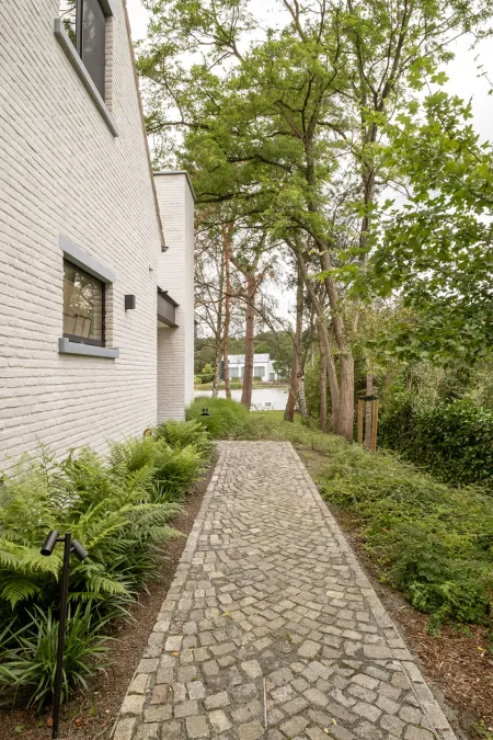 Kasseipad langs de witgekaleide villa met varens, omringd door bomen en zicht op het water