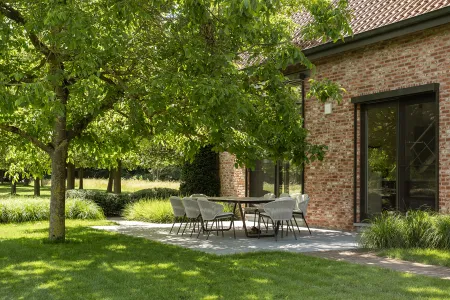 Lange eettafel met acht stoelen onder beschaduwende bomen, naast de glazen gevel van de hoeve.