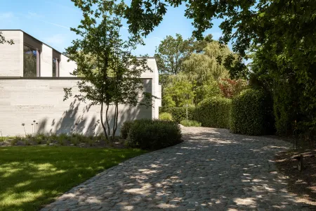 Glooiende kasseioprit tussen strakke hagen, omzoomd door bomen richting de villa-ingang.