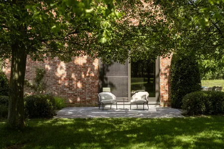 Twee loungestoelen op een schaduwrijk terras tegen de oude bakstenen gevel van de hoeve.