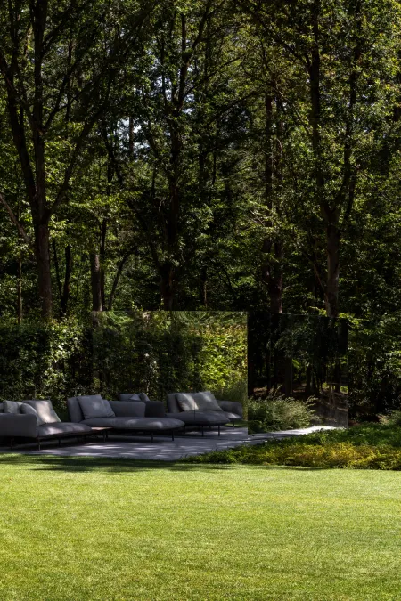 Loungezetel op een terras bij een spiegelende tuinwand, met dicht bos op de achtergrond.