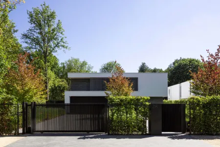 Zwart toegangshek met huisnummer, verscholen tussen dichte begroeiing en hoog bos.