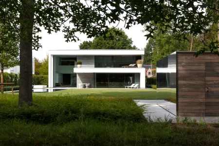 Zicht op de villa en houten tuinberging vanuit het gazon, met bomen die de tuin omkaderen.