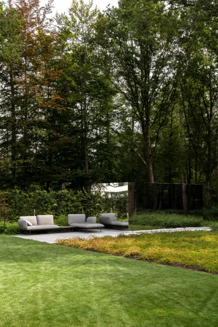 Loungezetel op een terras bij een spiegelende tuinwand, met dicht bos op de achtergrond.